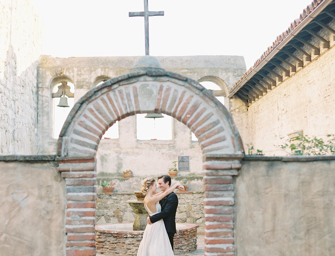 bride and groom hugging in front of Mission San Juan Capistrano courtyard