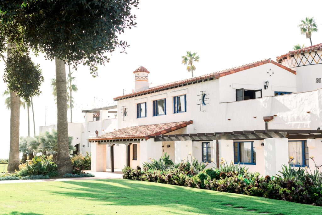 A white stucco building with red tile roof, wooden shutters, and arched windows, surrounded by green lawn, palm trees, and lush landscaping on a sunny day.