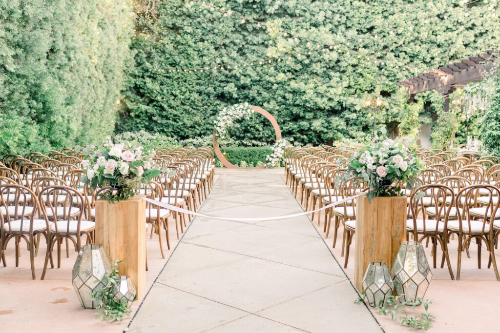 Outdoor wedding aisle with rows of wooden chairs, floral arrangements, glass lanterns, and a circular floral arch at the front, surrounded by lush greenery.