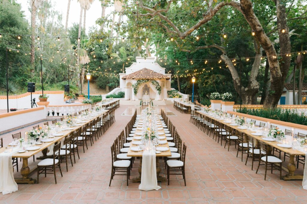 Outdoor wedding reception setup with two long banquet tables, white chairs, floral centerpieces, string lights overhead, and a decorated gazebo in the background surrounded by trees.