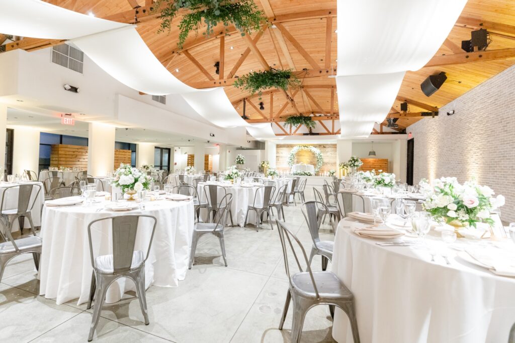 Bright, elegant banquet hall with round tables covered in white linens, silver chairs, white floral centerpieces, and draped white fabric on a wooden ceiling decorated with greenery.