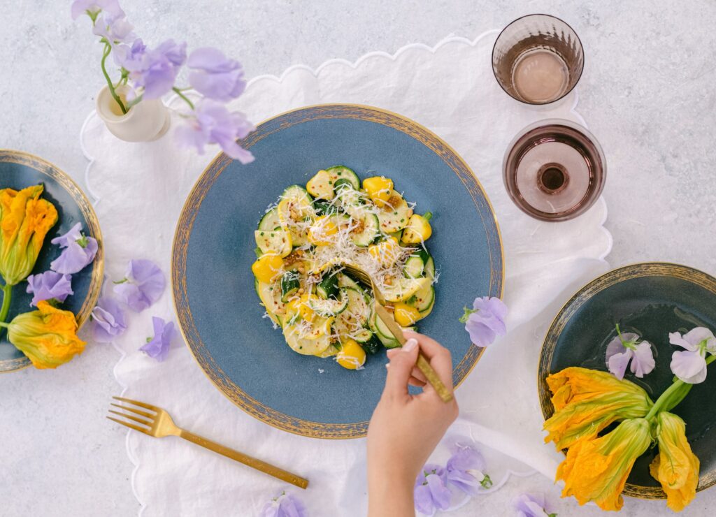 A hand holds a fork over a blue plate of pasta with yellow and green zucchini, garnished with cheese. Surrounding plates and flowers decorate a white tablecloth, with glasses and gold utensils nearby.