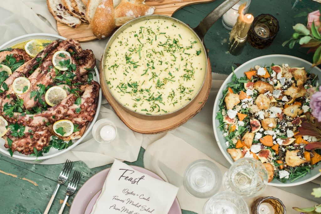 A table set for a meal features mashed potatoes, grilled octopus with lemon, salad with croutons and cheese, bread, drinks, candles, and a handwritten menu atop a green table with a beige table runner.