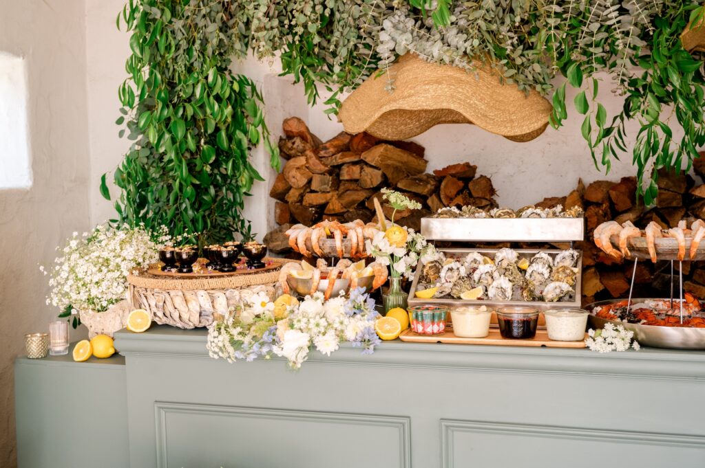 A seafood buffet with oysters, shrimp, and sauces is arranged on a green counter, decorated with flowers, greenery, and sliced lemons. Wood logs and hanging foliage fill the background.
