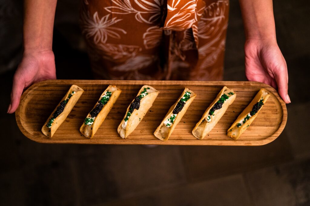A person holds a wooden tray with six neatly arranged, small rolled appetizers, each topped with cream and a dark garnish, against a dark background.