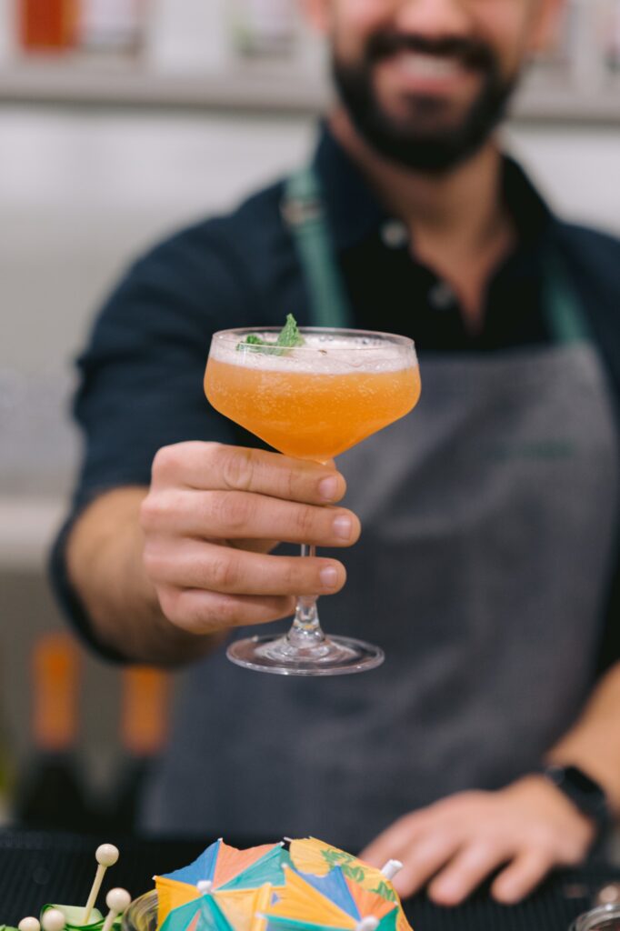 A bartender in a gray apron holds out an orange cocktail in a coupe glass, garnished with a mint leaf. A colorful cocktail umbrella is visible on the bar in the foreground.