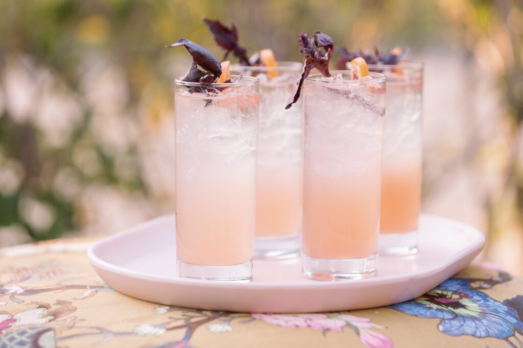 Four tall glasses of a pale pink cocktail with ice, each garnished with a sprig of purple basil and a twist of orange peel, arranged on a white tray outdoors.
