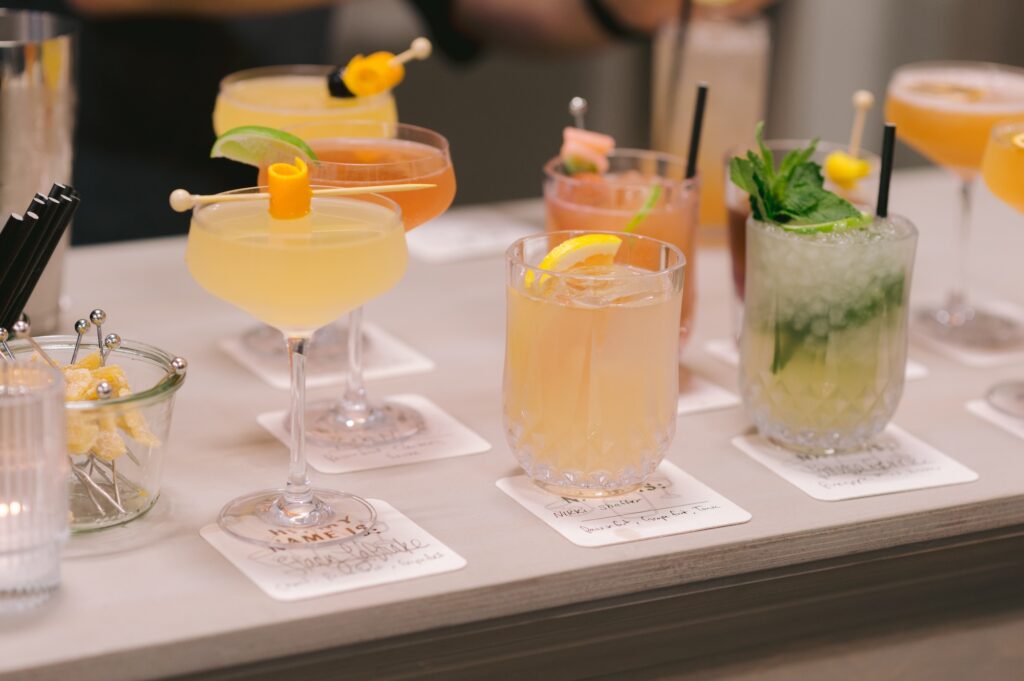 A row of assorted colorful cocktails in elegant glasses garnished with fruit and herbs, displayed on a bar counter with coasters and a bowl of sugar sticks.