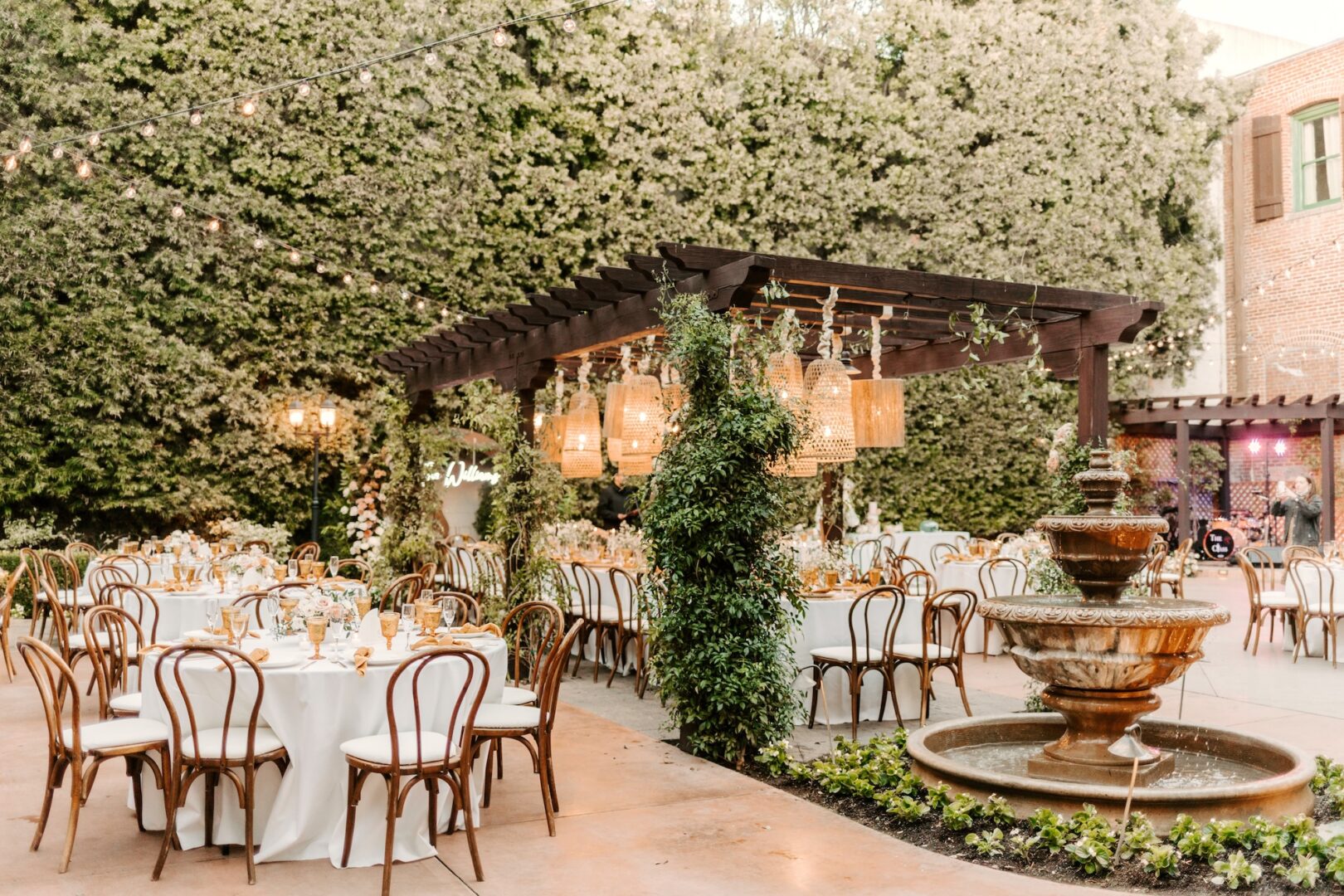 Outdoor wedding reception with round tables, wooden chairs, and a pergola decorated with hanging lights and greenery. A tiered fountain is in the foreground, surrounded by lush hedges and brick walls.