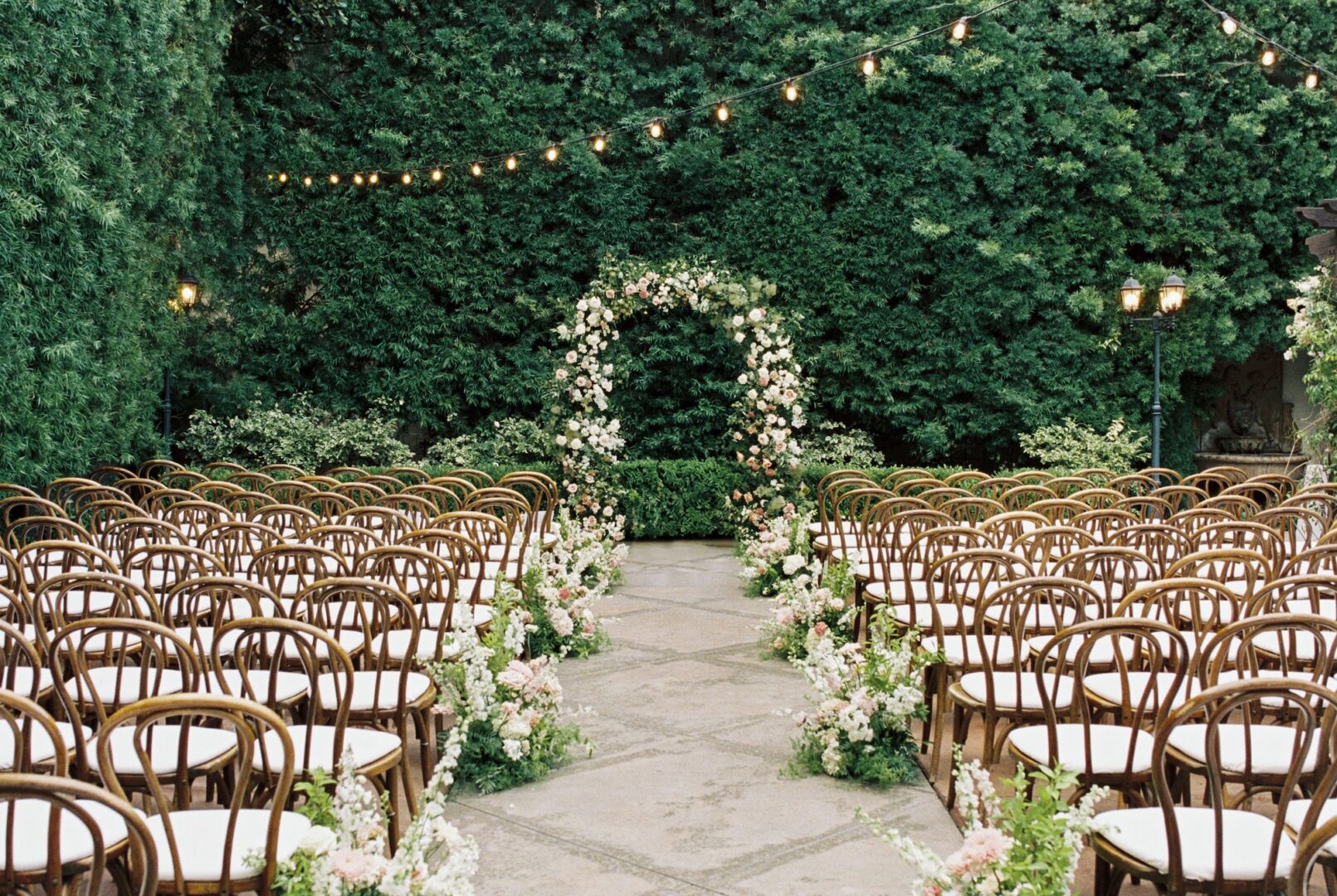 Rows of wooden chairs with white cushions face a flower-decorated archway outdoors, surrounded by lush greenery and string lights, ready for a wedding ceremony.