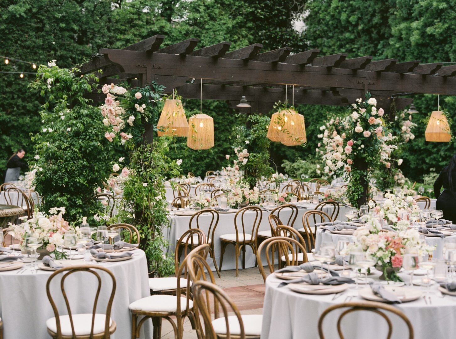 Outdoor garden wedding reception with round tables covered in white cloths, wooden chairs, floral centerpieces, and a pergola decorated with flowers and hanging lanterns. Lush green trees surround the elegant setting.