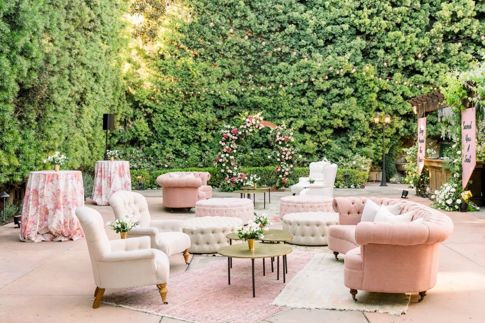 Elegant outdoor lounge area with pink and white tufted sofas, armchairs, round tables with flowers, floral arch, greenery backdrop, and string lights, creating a cozy, inviting atmosphere for an event or celebration.