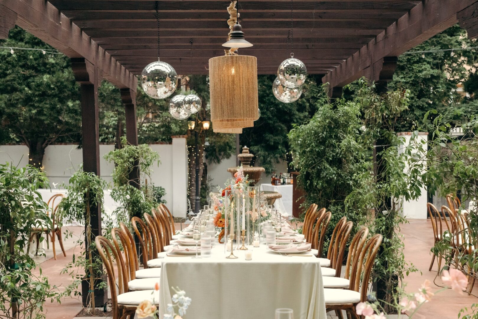 An elegant outdoor dining table set for a formal event under a pergola, with string lights, disco balls, wooden chairs, and lush greenery surrounding the area.