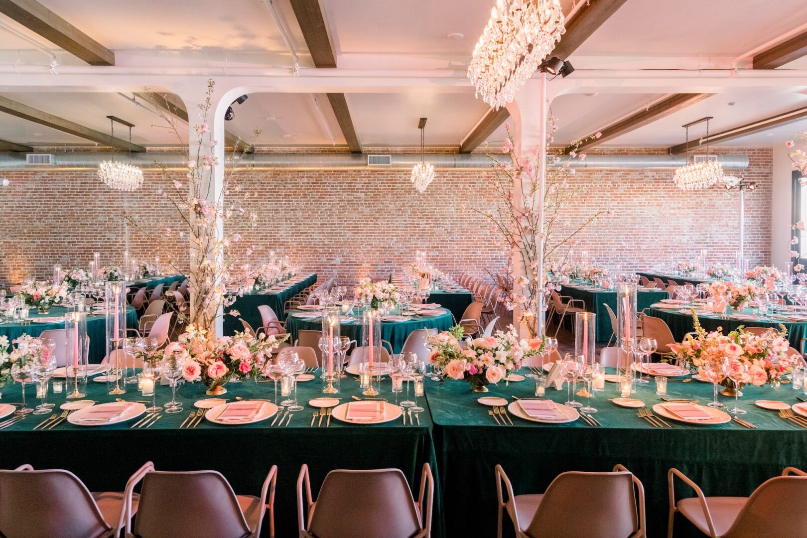 Elegant banquet hall with long tables covered in dark green cloth, adorned with pink and white floral arrangements, candles, glassware, and plates, set for a formal event under sparkling chandeliers.