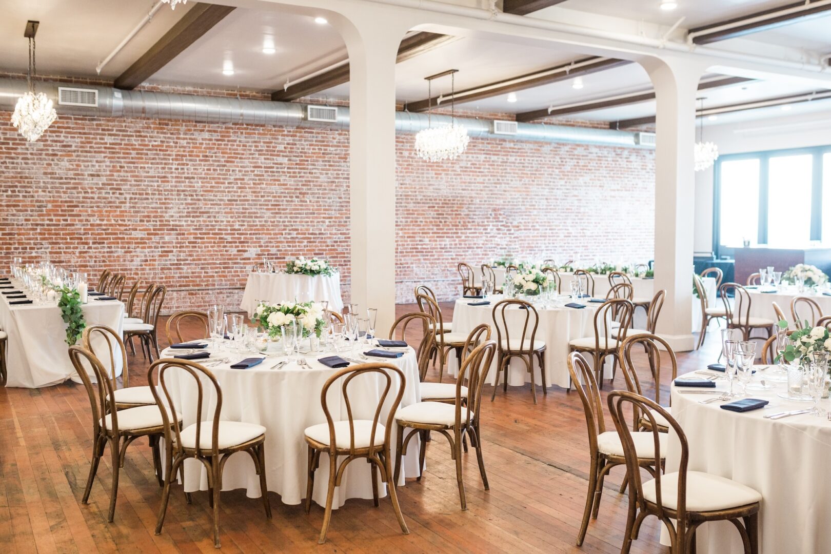 Elegant event space with round tables covered in white cloths, wooden chairs, floral centerpieces, and chandeliers, set in a room with exposed brick walls and wooden floors.