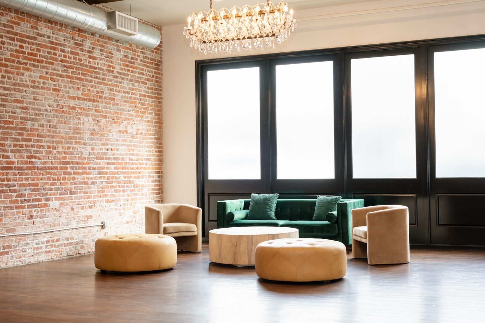 A modern lounge with a green velvet sofa, two beige armchairs, round beige ottomans, a circular table, a brick wall, large windows, and a chandelier overhead.