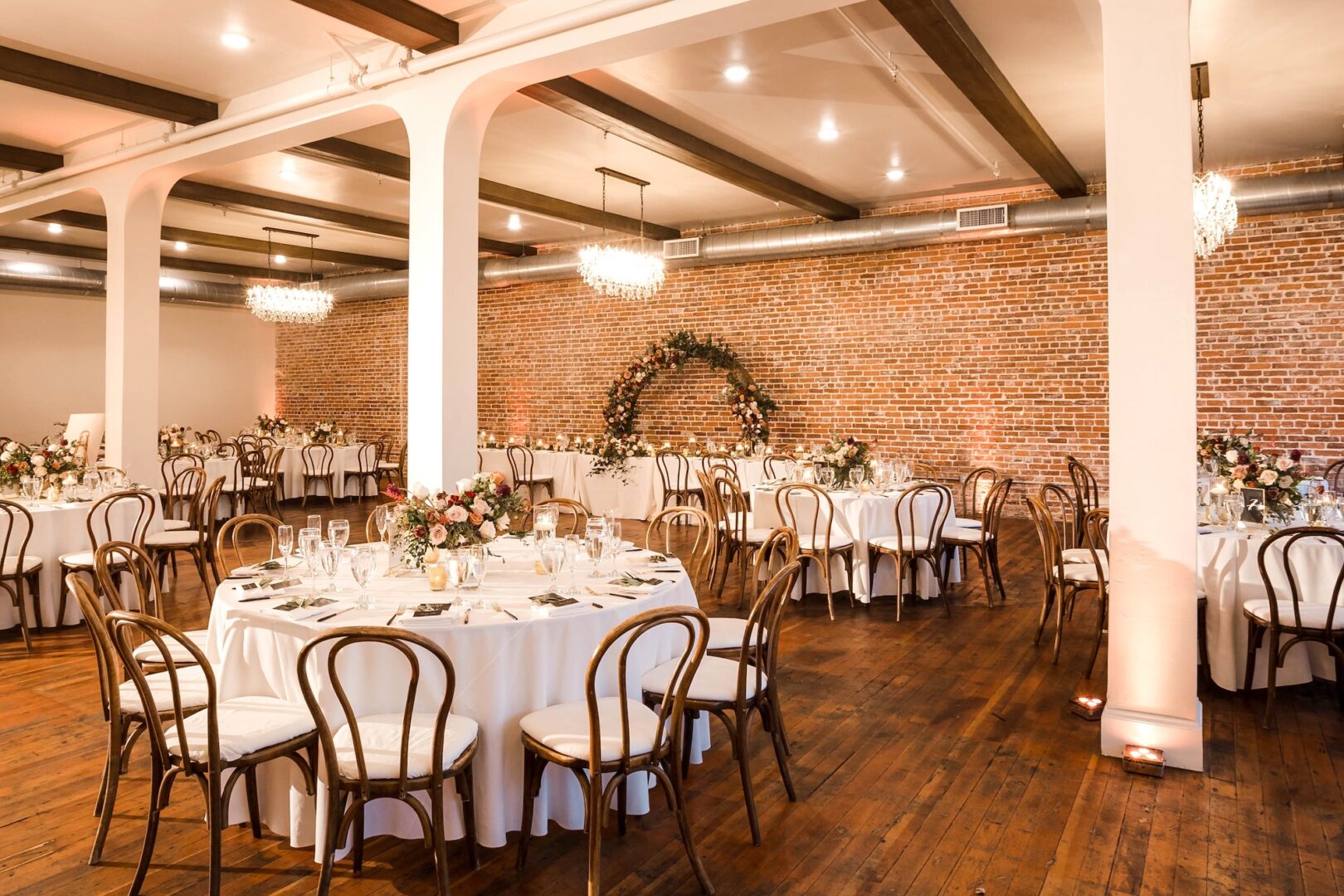 Elegant banquet hall with round tables covered in white cloths, floral centerpieces, wooden chairs, chandeliers, exposed brick walls, and a floral arch in the background, set up for a formal event.