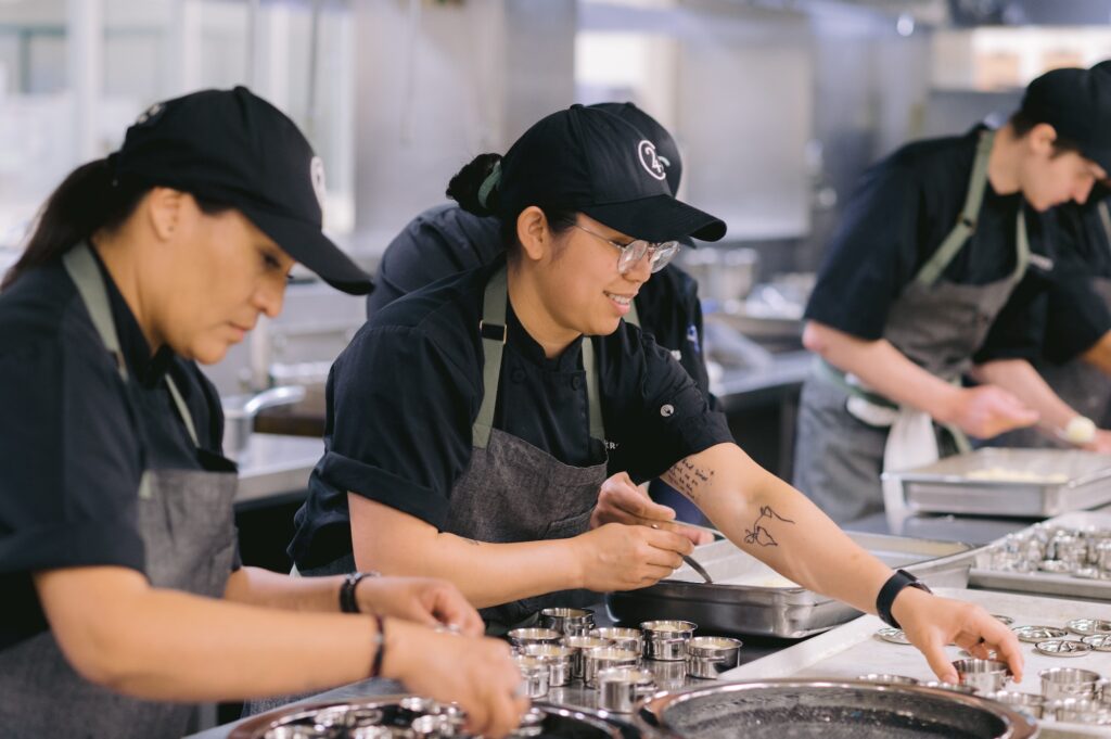 Three people in chef uniforms work together in a commercial kitchen, placing ingredients into metal containers. The focus is on the middle person, who is smiling and wearing glasses, a cap, and an apron.