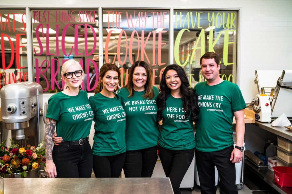 Five people in matching green T-shirts with food-related slogans stand together in a kitchen, smiling for the camera. Colorful slogans decorate the glass panels behind them.