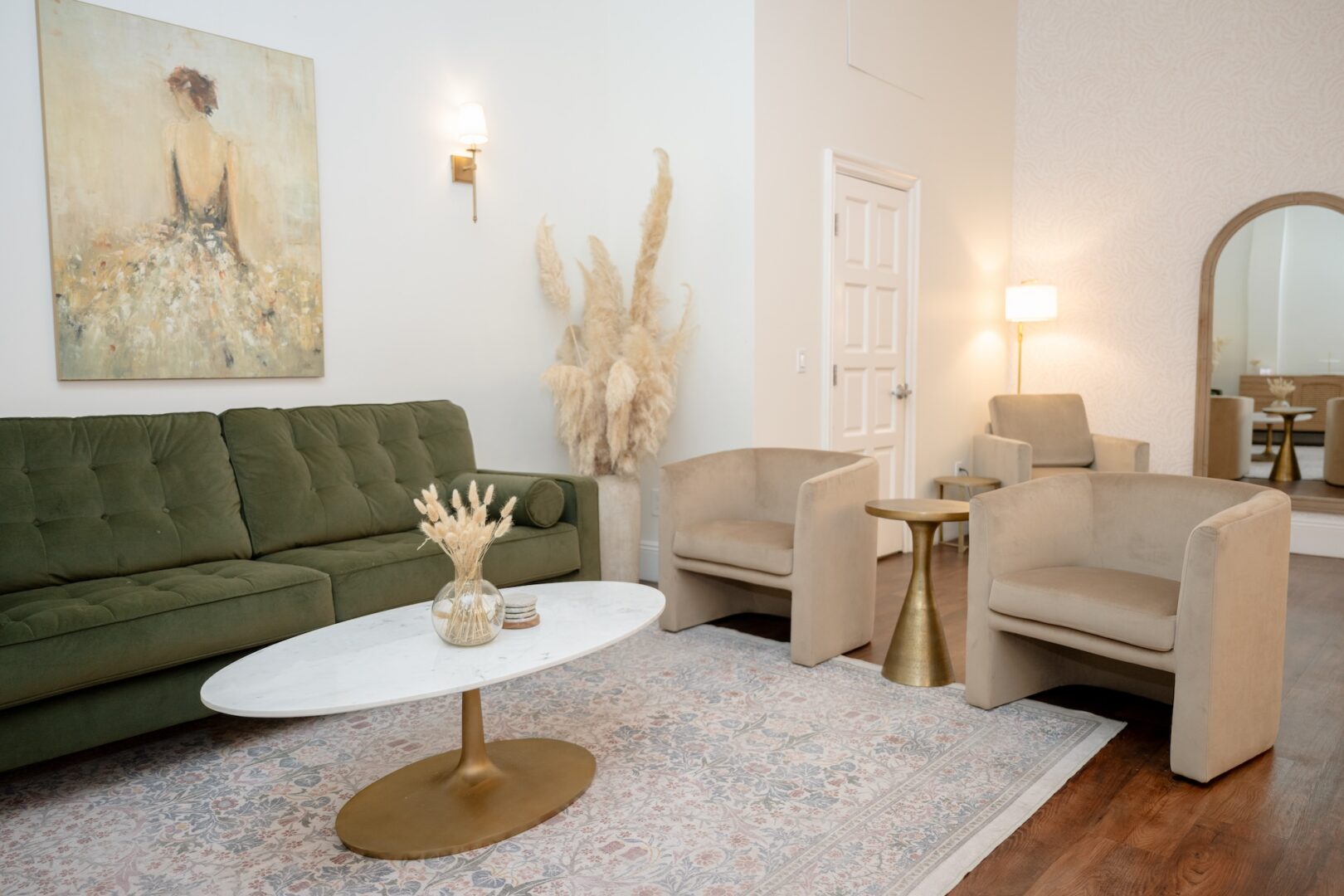 Modern living room with a green sofa, two beige chairs, a white oval coffee table, gold accents, pampas grass, a floor lamp, and a large wall mirror. Soft, neutral décor creates a cozy and elegant atmosphere.