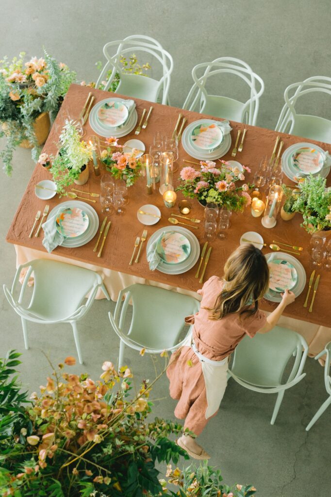A woman sets a table decorated with flowers, candles, gold cutlery, and pastel plates, viewed from above. The table is covered with a rust-colored cloth and surrounded by white chairs.