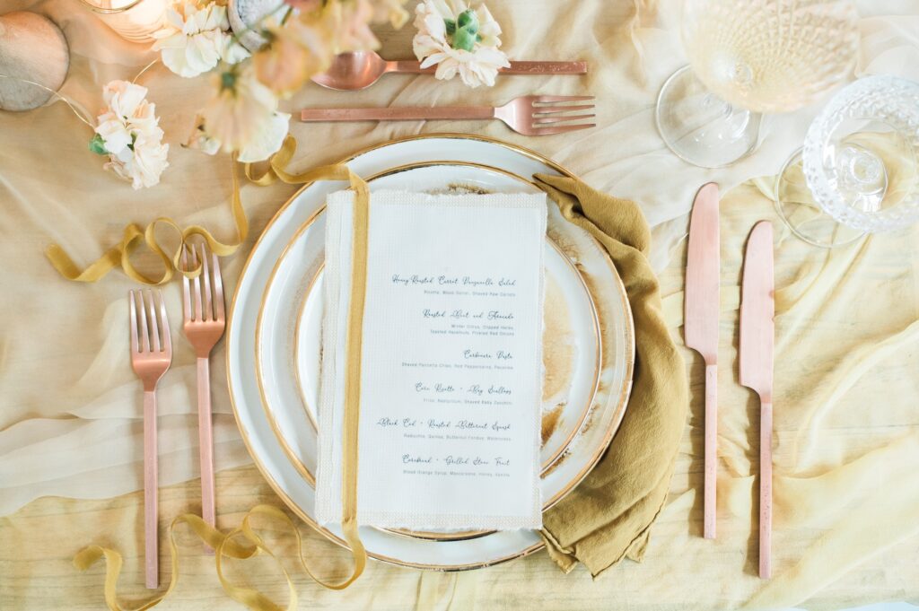 Elegant table setting with gold and blush pink cutlery, layered plates, a cloth menu, glassware, and soft floral accents on a yellow and cream tablecloth.